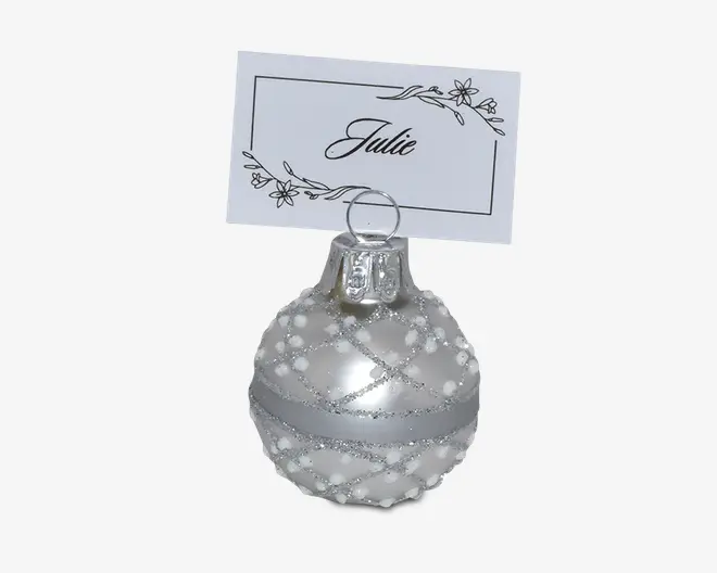 Rundt, sølvfarvet ornament med blank overflade, dekoreret med et krydsmønster af glimmer og små hvide perler. Ornamentet har en metalhætte foroven med en trådløkke til at holde et bordkort. Bordkortholderen er integreret i ornamentet, hvilket gør det velegnet til festlige bordopdækninger eller arrangementer.