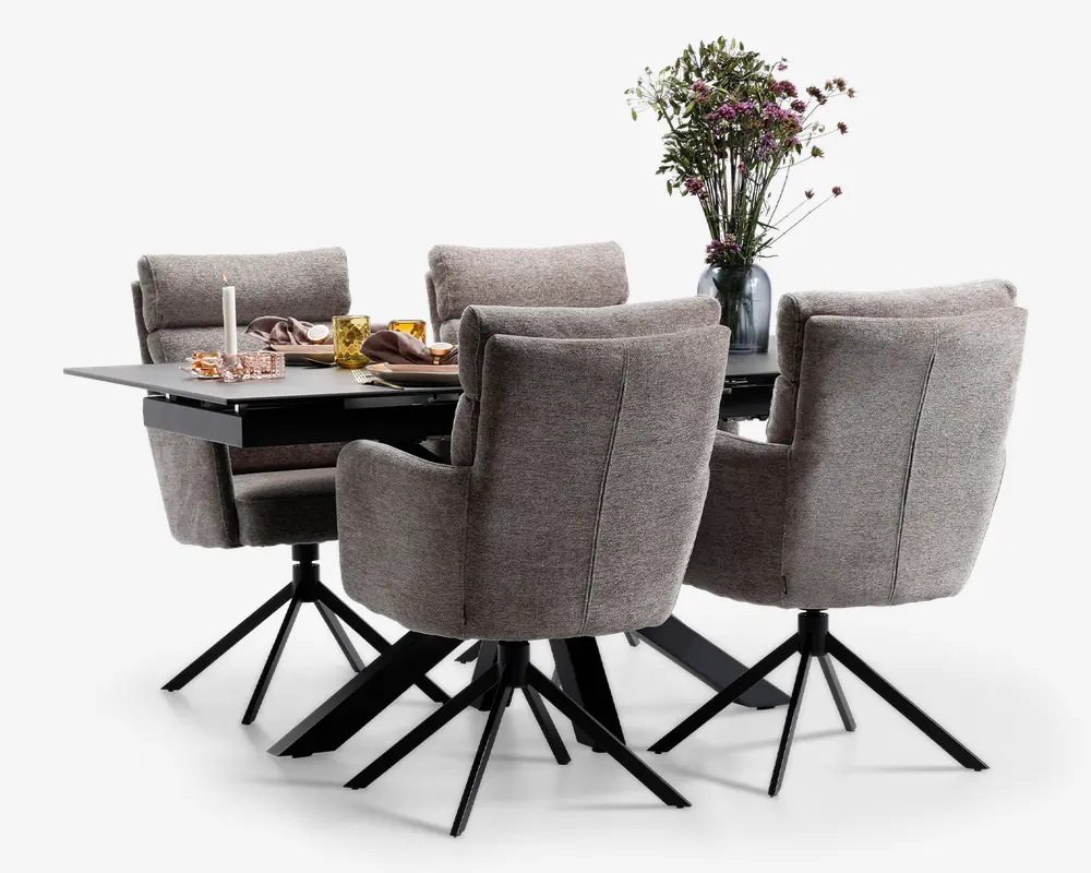 Et moderne spisesæt bestående af et rektangulært sort bord med mat overflade og seks polstrede stole. Stolene er betrukket med et blødt, lysegråt stof, har høje rygge med diskrete horisontale syninger, polstrede armlæn og står på sorte metalstel med fire ben og drejefunktion. Bordet er dækket med dekorative elementer såsom en vase med friske blomster, et stearinlys, glas og servietter, hvilket skaber en indbydende og elegant spiseatmosfære.