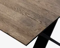 Nærbillede af en bordplade med naturlig træstruktur og let skrå kant. Overfladen viser forskellige træmønstre og knaster, hvilket giver et rustikt og autentisk udseende. Bordet understøttes af et solidt, sort metalben med et moderne, vinklet design.