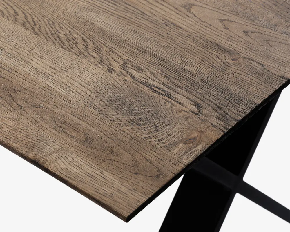 Nærbillede af en bordplade med naturlig træstruktur og let skrå kant. Overfladen viser forskellige træmønstre og knaster, hvilket giver et rustikt og autentisk udseende. Bordet understøttes af et solidt, sort metalben med et moderne, vinklet design.