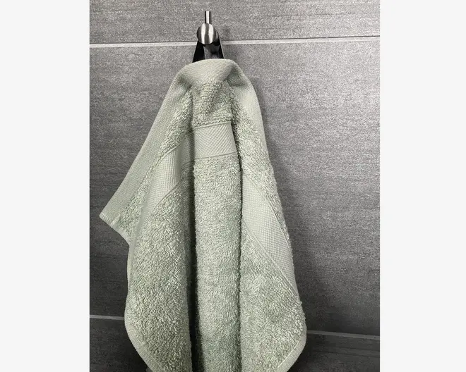 Et lysegrønt håndklæde hænger på en krog mod en grå flisevæg. Håndklædet har en blød, tekstureret overflade med en dekorativ kant, der indeholder et diskret geometrisk mønster. Kanterne er pænt syet, og stoffet ser tykt og absorberende ud, velegnet til daglig brug.