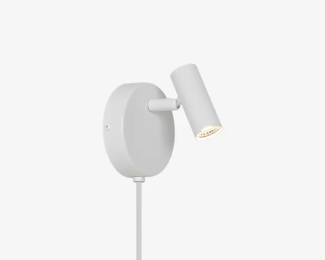 Hvid vægmonteret spotlampe med oval bagplade, kort justerbar arm og cylindrisk lampehoved vinklet nedad; en slank lodret støtte går ned fra basen.
