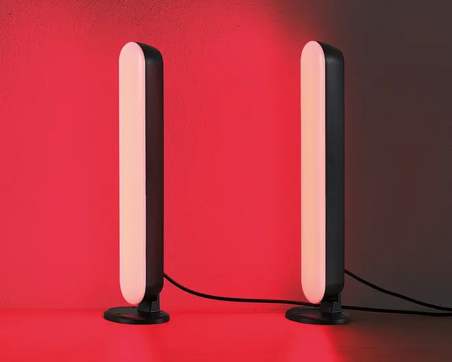 Et par slanke, lodrette LED-lysstænger med afrundede kanter, monteret på små, runde sorte baser. Lysene udsender en blød rød glød, der oplyser området omkring dem. Hver stang er forbundet med en synlig sort strømkabel. Designet er moderne og minimalistisk, velegnet til stemnings- eller accentbelysning.