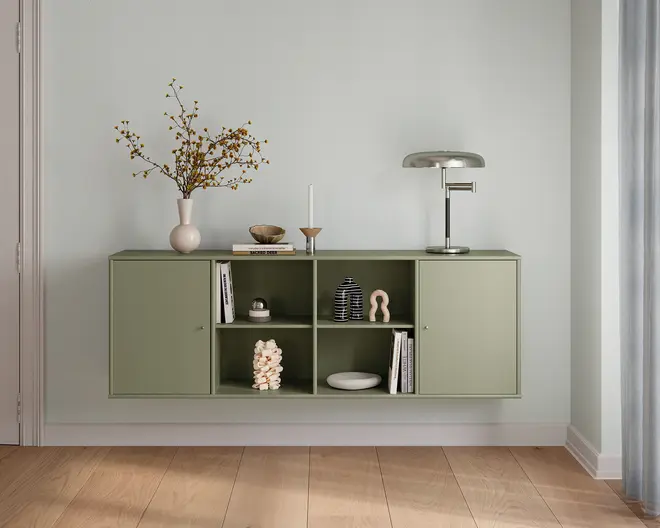 Vægmonteret opbevaringsenhed i en afdæmpet salviegrøn farve med et minimalistisk design. Enheden har to lukkede skabe i siderne og fire åbne hylder i midten. Hylderne viser dekorative genstande som en hvid vase med grene, en metalbordlampe, en lysestage, en lille skål, bøger, en skulpturel figur og en stak hvid koral. Det samlede udtryk er moderne og organiseret med en blød, mat finish og rene linjer.