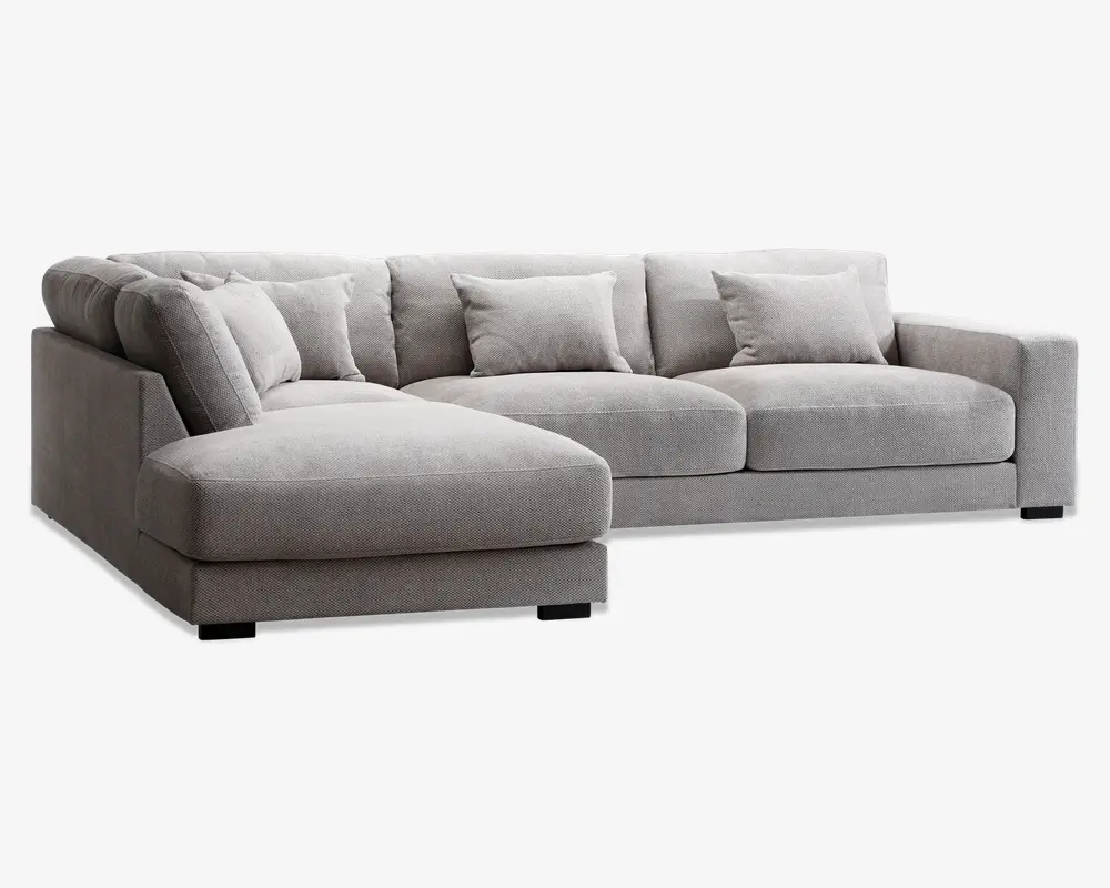 Moderne hjørnesofa i lysegråt stof med venstrevendt chaiselong. Sofaen har brede, firkantede armlæn, dybe sædehynder og fem matchende ryghynder for ekstra komfort. Betrækket har en diskret tekstureret vævning, og sofaen står på lave, sorte blokformede ben.