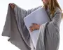 En person bærer et lysegråt, oversized tæppe med brede ærmer og blød, quiltet tekstur. Tæppet falder løst og ser ud til at være lavet af et blødt, hyggeligt materiale. Personen holder en stor, rektangulær, hvid pude med afrundede hjørner, som ser støttende og behagelig ud. Det samlede indtryk er varme og komfort, ideelt til afslapning derhjemme.