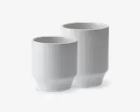 To minimalistiske, cylindriske hvide kopper med mat finish. Begge kopper har en let tilspidset bund og en glat, enkel overflade, hvilket giver dem et moderne og elegant udtryk. Den ene kop er højere end den anden, hvilket viser, at de fås i forskellige størrelser.