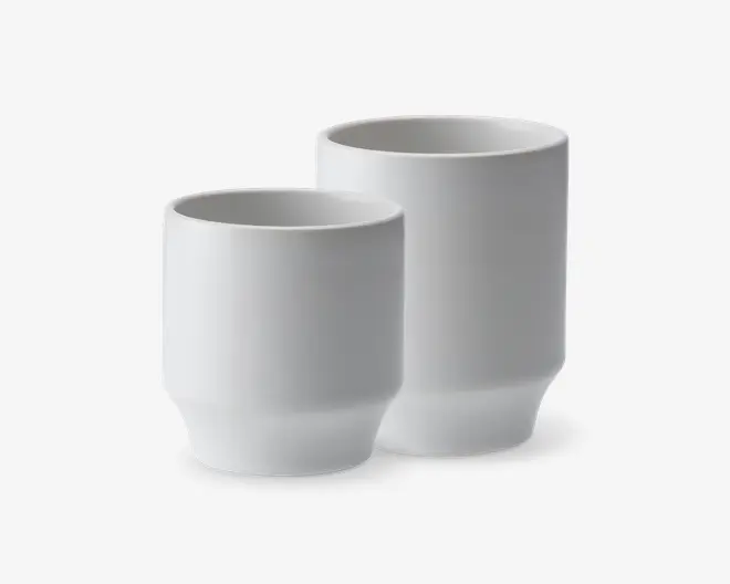 To minimalistiske, cylindriske hvide kopper med mat finish. Begge kopper har en let tilspidset bund og en glat, enkel overflade, hvilket giver dem et moderne og elegant udtryk. Den ene kop er højere end den anden, hvilket viser, at de fås i forskellige størrelser.