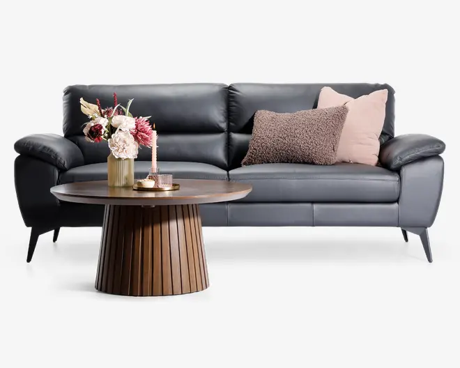Et moderne, rundt sofabord med rillet træfod og glat, cirkulær bordplade. Bordet er dekoreret med en guldfarvet vase med kunstige blomster i nuancer af pink, creme og bordeaux, et lyserødt dekorativt stearinlys og et lille fad med to glas fyrfadsstager. Bordet står foran en mørkegrå lædersofa med bløde, skrå armlæn og to pyntepuder – én i blød rosa og én i tekstureret, brunligt stof.