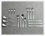 Et sæt bestik i rustfrit stål arrangeret på en grå overflade, inklusive tre store spiseskeer, tre små teskeer, tre gafler, tre knive, én suppeske og én dessertske. Bestikket har et enkelt, moderne design med glatte, afrundede håndtag og en blank finish.