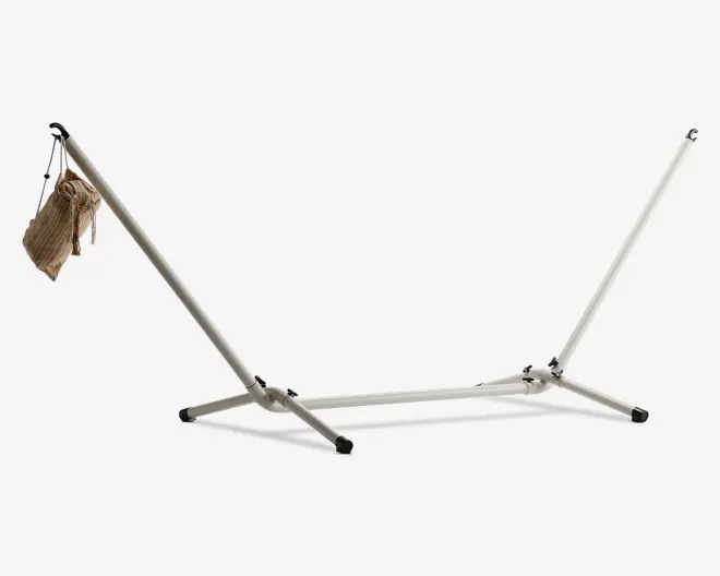 Solid hængekøjestativ i metal med pulverlakeret overflade, udstyret med fire bredt placerede ben for stabilitet og gummifødder for at forhindre glidning. Stativet har skrå arme med kroge til fastgørelse af en hængekøje, og en sammenfoldet stofhængekøje hænger i den ene ende, hvilket viser, at den er nem at opbevare og transportere.