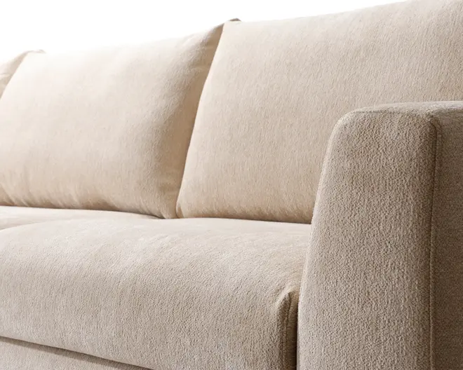 Nærbillede af en sofa med blødt, tekstureret beige betræk. Sæde- og ryghynderne er fyldige og godt polstrede, hvilket giver behagelig støtte. Armlænet er bredt og let afrundet, og matcher det minimalistiske og moderne design. Stoffet virker slidstærkt og nemt at rengøre.