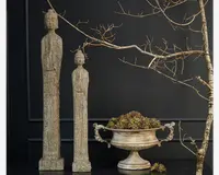 To høje, slanke dekorative statuer med rustik, patineret stenfinish står side om side. Hver statue har aflange kroppe og enkle, stiliserede detaljer. Ved siden af står en bred sølvfarvet urne med dekorative håndtag, fyldt med grønne og brune sukkulenter. Baggrunden består af en mørk væg og en nøgen gren, hvilket giver scenen et naturligt og elegant udtryk.