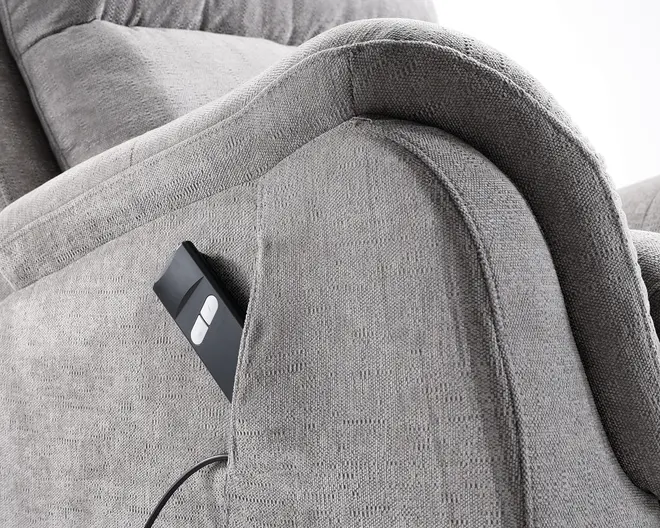 Nærbillede af et armlæn betrukket med lysegråt stof, der har en indbygget sidelomme. Lommen indeholder en sort fjernbetjening med to hvide knapper, hvilket tyder på, at møblet har en elektrisk eller motoriseret funktion. Stoffet har en blød, tekstureret overflade, og armlænet er tykt polstret for ekstra komfort.