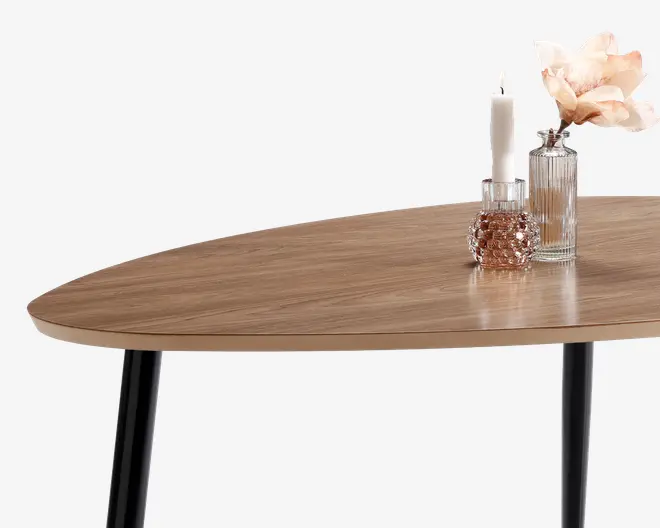 Billedet viser en bordplade med en glat, lys træfinish og afrundede kanter. Bordet har slanke, sorte metalben. På bordet står dekorative genstande, herunder en rillet glasvase med en lyserød kunstig blomst, et højt hvidt stearinlys i en klar glaslysestage og en lille, rund, tekstureret glaskrukke.