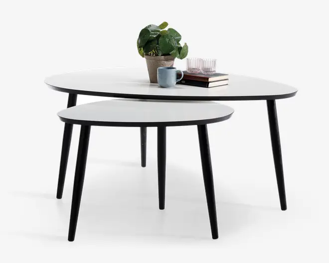 Sæt med to indskudsborde med runde, hvide bordplader og slanke, sorte skrå ben. Det større bord er dekoreret med en potteplante, et blåt krus, to glas og en stak bøger, mens det mindre bord er uden pynt. Bordene har et minimalistisk, moderne design og kan placeres sammen eller hver for sig.