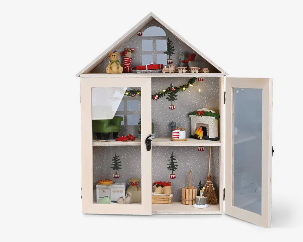 Et miniature vitrineskab formet som et hus med to glasdøre, dekoreret til jul. Indvendigt er der tre hylder med små møbler og festlige tilbehør, herunder en grøn lænestol, en pejs med julestrømper, en guirlande med lys, et lille køkkenområde og forskellige julepynt som en bamse, en slæde og indpakkede gaver. Baggrunden har snefnug og juletræer, hvilket understreger det hyggelige juletema.