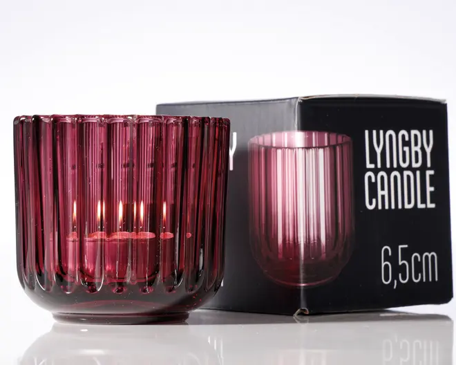 En lille, rund lysestage lavet af dybrødt glas med lodrette riller. Glasset er gennemsigtigt, så lyset fra stearinlyset skinner igennem og skaber en varm glød. Lysestagen står ved siden af sin emballage, som viser et billede af produktet og angiver højden til 6,5 cm.