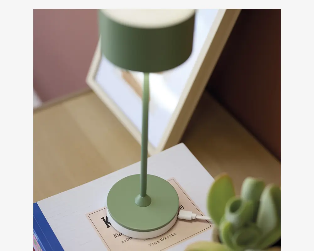 En moderne bordlampe med mat grøn finish, der har en slank cylindrisk stang og en rund fod. Lampen har et enkelt, minimalistisk design med rene linjer. En hvid opladerkabel er tilsluttet lampens fod, hvilket indikerer, at den muligvis er genopladelig eller USB-drevet. Lampen står på en lys overflade ved siden af en bog og en lille plante.
