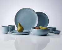 Et sæt service i en blød blå farve med blank overflade, bestående af store og små tallerkener, skåle, krus og kopper. Delene har et enkelt, moderne design med let afrundede kanter og en diskret, håndlavet struktur. Sættet er vist med frugt for at illustrere størrelse og anvendelse.
