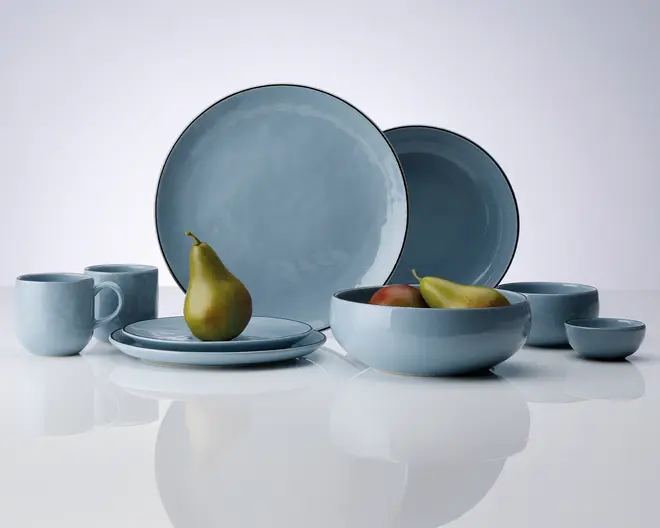 Et sæt service i en blød blå farve med blank overflade, bestående af store og små tallerkener, skåle, krus og kopper. Delene har et enkelt, moderne design med let afrundede kanter og en diskret, håndlavet struktur. Sættet er vist med frugt for at illustrere størrelse og anvendelse.