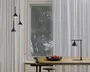 Minimalistisk interiør med et træbord, hvorpå der står en mat sort skål med tre røde æbler. Bordet har en elegant sort metalbordlampe med konisk skærm. Tre matchende pendellamper med kegleformede skærme hænger fra loftet. Baggrunden består af hvide gardiner fra gulv til loft og et stort vindue med udsigt til et træ udenfor.