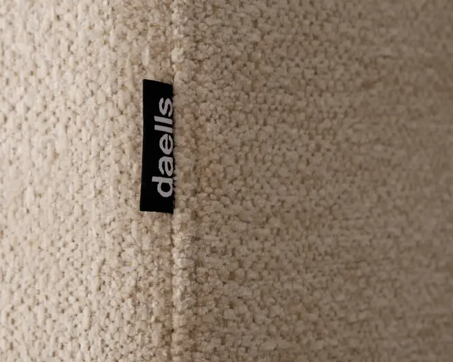 Nærbillede af et tekstureret, beige stof med en blød, løkket bouclé-overflade. Stoffet er syet sammen ved en syning, hvor der sidder et lille sort stofmærke med mærket 'daells' i hvide bogstaver. Materialet fremstår blødt og hyggeligt, velegnet til polstring eller møbler.