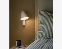 En hvid vægmonteret lampe med en konisk skærm, der udsender et varmt lys. Lampen har et minimalistisk design og er placeret ved siden af en seng med stribet sengetøj. Lampens skærm leder lyset nedad, hvilket gør den velegnet til læsning eller stemningsbelysning.