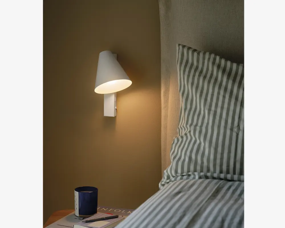 En hvid vægmonteret lampe med en konisk skærm, der udsender et varmt lys. Lampen har et minimalistisk design og er placeret ved siden af en seng med stribet sengetøj. Lampens skærm leder lyset nedad, hvilket gør den velegnet til læsning eller stemningsbelysning.