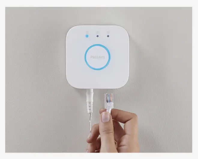 En hånd holder et Ethernet-kabel tæt på en lille, firkantet, hvid enhed, der er monteret på en væg. Enheden har afrundede hjørner, tre indikatorlamper øverst og en lysende blå ring omkring ordet 'Philips' i midten. Et andet kabel er allerede tilsluttet enheden. Enheden er designet til kablet netværksforbindelse og har et rent, minimalistisk udseende.