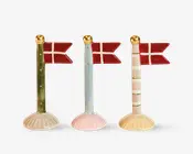 Tre dekorative flagstænger, hver med et rødt og hvidt flag med kors. Flagstængerne har forskellige designs: én er grøn med guldfarvede prikker, én er lyseblå med guldfarvede prikker, og én er hvid med lyserøde striber og grønne ringe. Hver stang har en guldfarvet kugle på toppen og står på en muslingeformet fod i matchende pastelfarver.