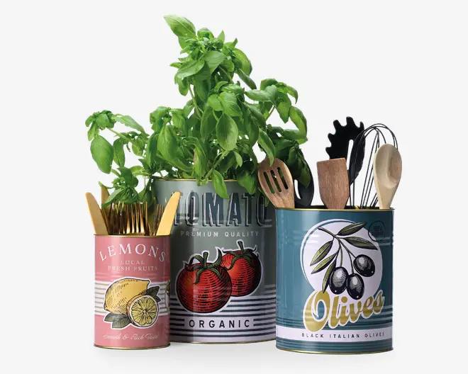 Tre dekorative metaldåser med vintage-inspirerede etiketter vises. Den mindste dåse, mærket med citroner, indeholder guldfarvet bestik. Den mellemstore dåse, mærket med tomater, indeholder en frodig grøn basilikumplante samt nogle træredskaber. Den største dåse, mærket med oliven, rummer forskellige køkkenredskaber, herunder træskeer og en pastaske. Hver dåse har farverigt, retro-inspireret design og tekst, hvilket giver dem et charmerende, rustikt køkkenudtryk.