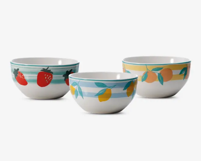 Sæt med tre keramiske skåle, hver med unikt håndmalet frugtmotiv og farverige vandrette striber. Én skål har røde jordbær med grønne blade på turkise striber, en anden har gule citroner med grønne blade på lyseblå striber, og den tredje viser orange ferskner med grønne blade på gule striber. Skålene har en blank overflade og rund form, velegnet til servering eller omrøring.
