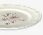 Rund keramiktallerken med let bølget kant og blank overflade. Midten har et fint blomsterdesign i bløde rosa, lilla og grønne nuancer, hvilket giver et klassisk og elegant udtryk. Kanten er let hævet og tilføjer et diskret dekorativt element.