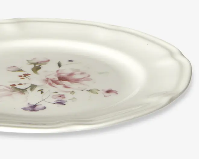 Rund keramiktallerken med let bølget kant og blank overflade. Midten har et fint blomsterdesign i bløde rosa, lilla og grønne nuancer, hvilket giver et klassisk og elegant udtryk. Kanten er let hævet og tilføjer et diskret dekorativt element.