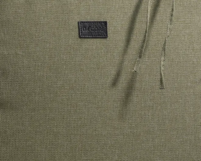 Nærbillede af et tekstureret olivengrønt stof med et diskret vævet mønster. Der er en lille, rektangulær sort mærkat med mærket 'DACORE' syet nær toppen i midten. To tynde snore er synlige i højre side, hvilket antyder en justerbar funktion.