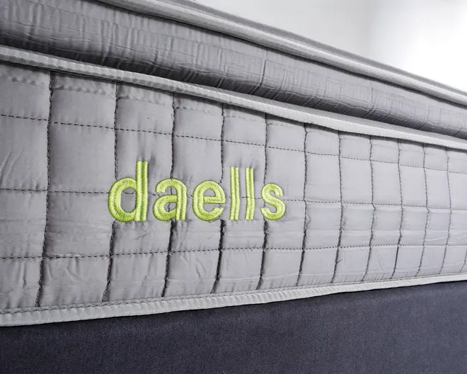Nærbillede af en madras' sidepanel med lysegråt, quiltet stof i et ternet mønster. Brandnavnet 'daells' er broderet med lysegrønt garn. Syninger og polstring er tydelige, hvilket indikerer en solid og støttende kant.