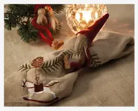 En beige linnedug med detaljeret broderi, der viser en hvid gyngehest med rød sadel og en hængende kogler-ornament. Servietten er rullet sammen med en festlig servietring formet som en lille nisse med rød spids hue, langt hvidt skæg og næse og hænder af træ. I baggrunden ses en glaskrone med levende lys, der spreder et varmt skær, samt nogle grangrene for en hyggelig, julet stemning.