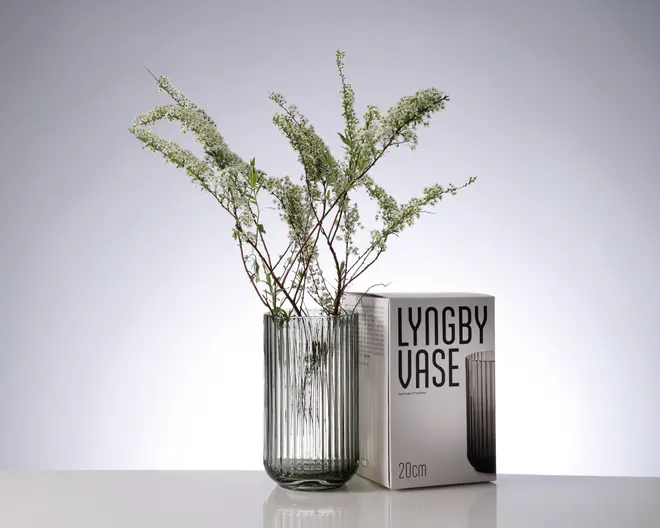En klar, cylindrisk glasvase med lodrette riller, placeret ved siden af sin emballageæske. Vasens design er moderne og minimalistisk. Den er fyldt med fine grønne grene og små hvide blomster. På æsken står der 'Lyngby Vase', og det fremgår, at vasen er 20 cm høj.
