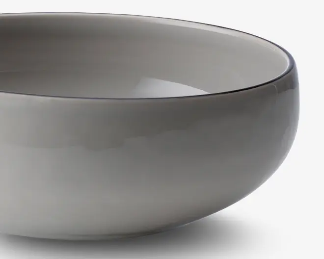 Et nærbillede af en glat, rund skål med en blank, lysegrå overflade. Skålen har et enkelt, minimalistisk design med blødt buede sider og en let tyk kant. Både indersiden og ydersiden har samme farve, hvilket giver et moderne og elegant udtryk.