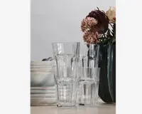 Fire klare glasdrikkeglas med facetslebet design, stablet oven i hinanden. Glassene har en let udvidet kant og en solid bund, velegnet til daglig brug. De står på et bord ved siden af en stak tallerkener og en mørk vase med kunstige blomster.
