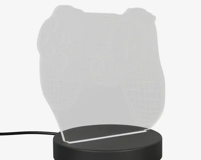 En dekorativ LED-lampe med en klar akrylplade formet som en videospil-controller, med indgraverede detaljer der fremhæver knapper og konturer. Pladen er monteret på en rund, sort base, som indeholder LED-lyskilden og har et strømkabel, der går ud fra siden. Lampen er designet til at skabe en 3D-effekt, når den er tændt.
