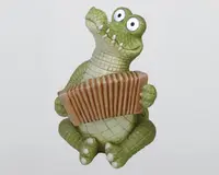 En finurlig figur af en grøn krokodille med overdrevne, tegneserieagtige træk, herunder store, udstående øjne og et bredt, tandet smil. Krokodillen sidder oprejst og holder et brunt harmonika med begge hænder. Kroppens og lemmernes overflade er tekstureret, så den ligner skæl, og tæerne har hvide spidser. Stilen er legende og humoristisk.