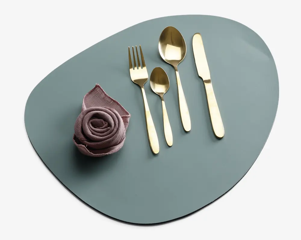 Et sæt med fire guldfarvede bestikdele, herunder en gaffel, en lille ske, en stor ske og en kniv, arrangeret pænt på en mat, uregelmæssigt formet lyseblå dækkeserviet. Ved siden af bestikket ligger en støvet rosa stofserviet foldet som en rose, hvilket giver et elegant udtryk.