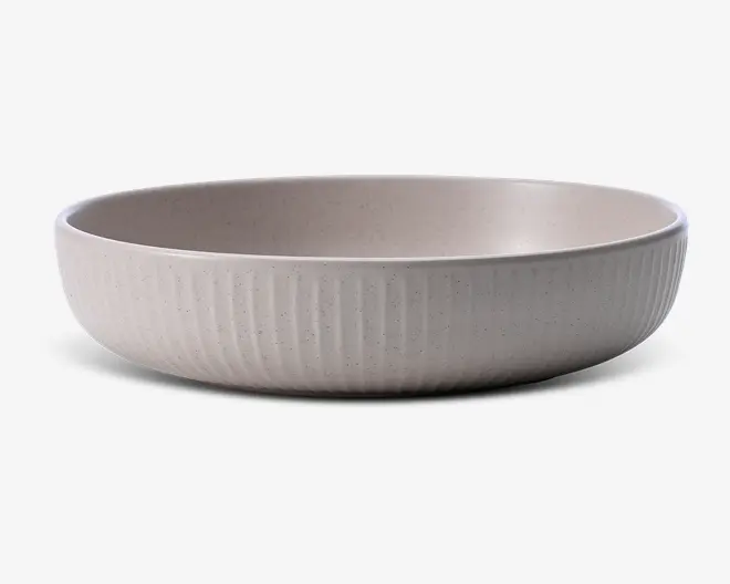 En lav, rund skål med mat, lysebeige overflade. Ydersiden har en diskret lodret rillet struktur, som giver både taktilt og visuelt udtryk. Indersiden er glat og uden dekoration, og skålen har et let plettet udseende, hvilket giver den et håndlavet, kunsthåndværkspræg.