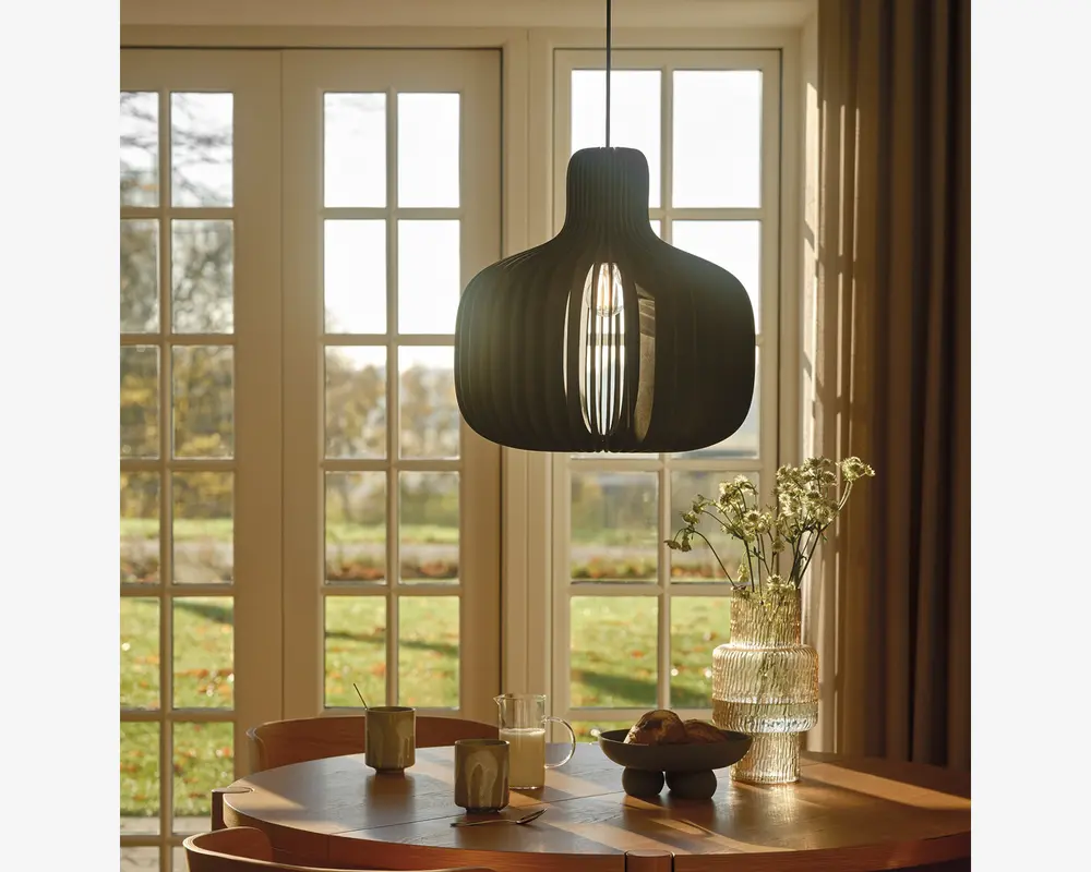Loftlampe med et moderne, skulpturelt design, der har en sort, buet skærm lavet af lameller, som lader lyset sive igennem og skaber et dekorativt mønster. Lampen hænger over et rundt spisebord i træ, hvorpå der står en glasvase med hvide blomster, en keramikskål, krus og en glaskande, alt sammen oplyst af naturligt lys fra store vinduer i baggrunden.