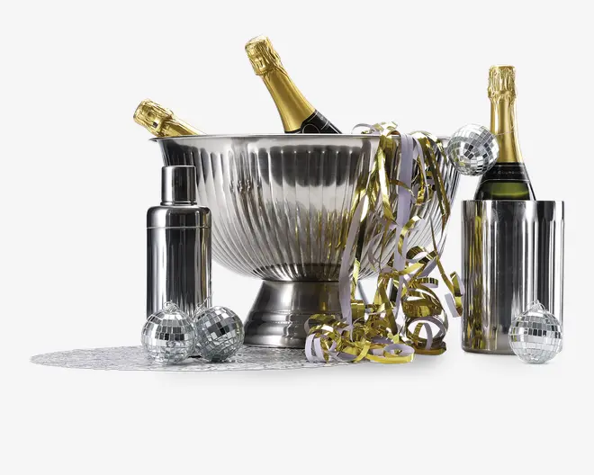 Billedet viser en festlig opstilling med en stor, riflede isspand i rustfrit stål, der indeholder flasker champagne. Rundt om spanden står en elegant metalcocktailshaker, en høj cylindrisk vinkøler og flere små diskokugledekorationer. Guld- og hvide krøllede bånd er draperet over spanden og spredt på bordet, hvilket understreger den festlige stemning. Udtrykket er elegant og klar til fest med fokus på metalliske overflader og glitrende detaljer.