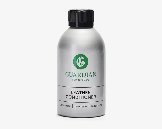 En sølvfarvet plastflaske med sort skruelåg, der har et grønt 'G'-logo og mærket 'Guardian Furniture Care' trykt i grønt. Etiketten viser også 'Leather Conditioner' med fed sort skrift, samt yderligere tekst på flere sprog nederst. Flasken har et enkelt, moderne design og ser ud til at være nem at holde og hælde af.