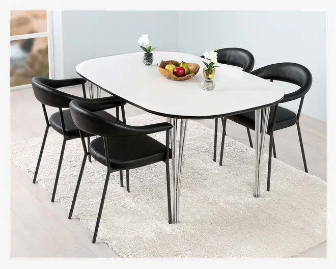 Moderne spisesæt med et hvidt, ovalt bord med slanke kromben og fire sorte stole med buede rygge og polstrede sæder. Bordet er pyntet med en træskål med frugt, herunder æbler og bananer, samt to små vaser med hvide blomster. Sættet står på et lyst, tekstureret tæppe i et lyst rum med lyse trægulve.