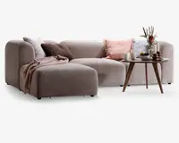 En moderne, L-formet sofa med blødt, lysebeige stofbetræk. Sofaen har afrundede armlæn og lav ryg, hvilket giver et hyggeligt og indbydende udtryk. Den er pyntet med flere dekorative puder i rosa, brun og grå nuancer samt et tekstureret tæppe lagt over chaiselongen. Foran sofaen står et rundt sofabord i træ med skrå ben, hvor der står en vase med blomster, stearinlys, kopper og en lille skål, hvilket tilfører varme og stil til rummet.